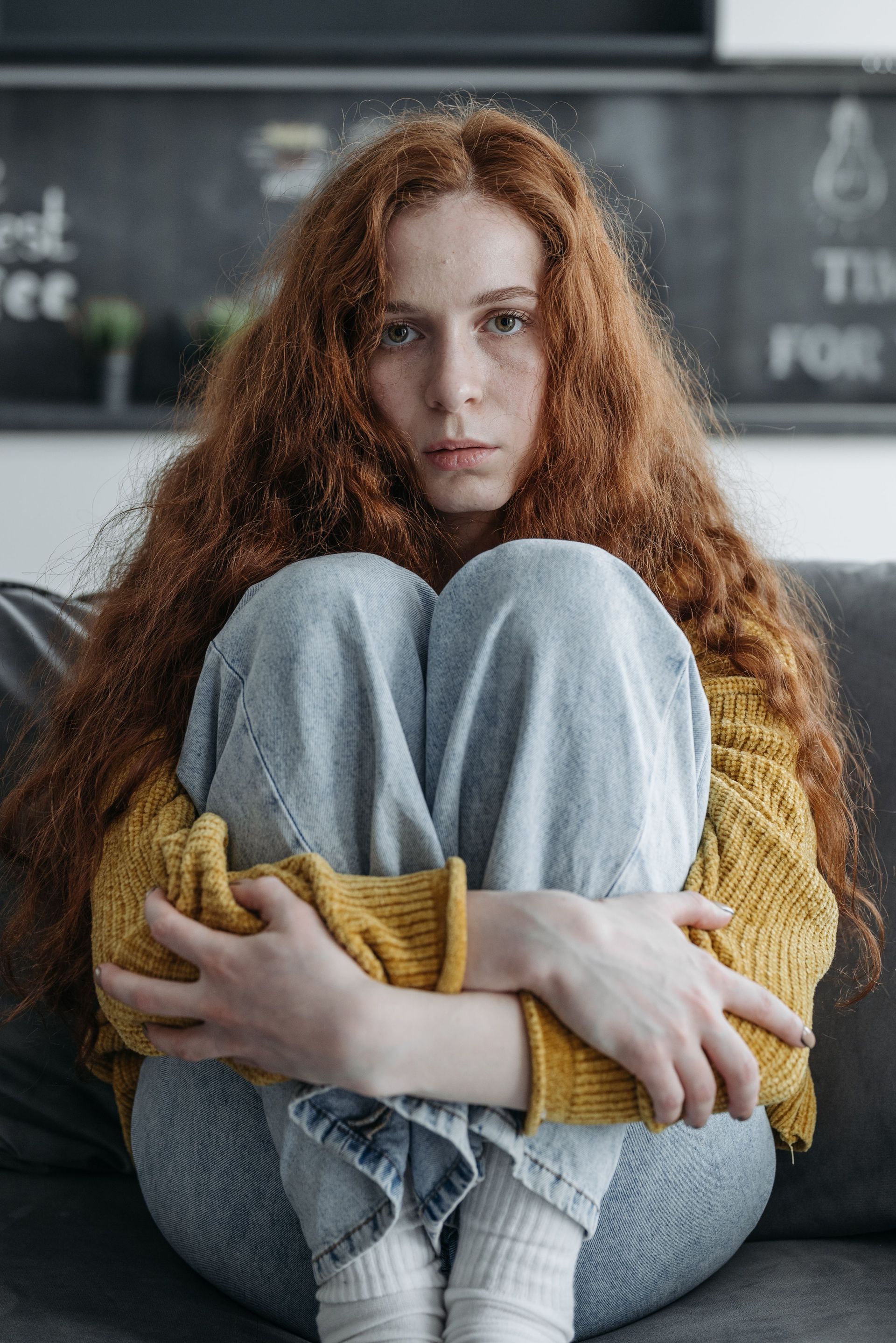 Curly-haired person sitting curled up on a sofa, arms around knees, wearing a yellow sweater and jeans.
