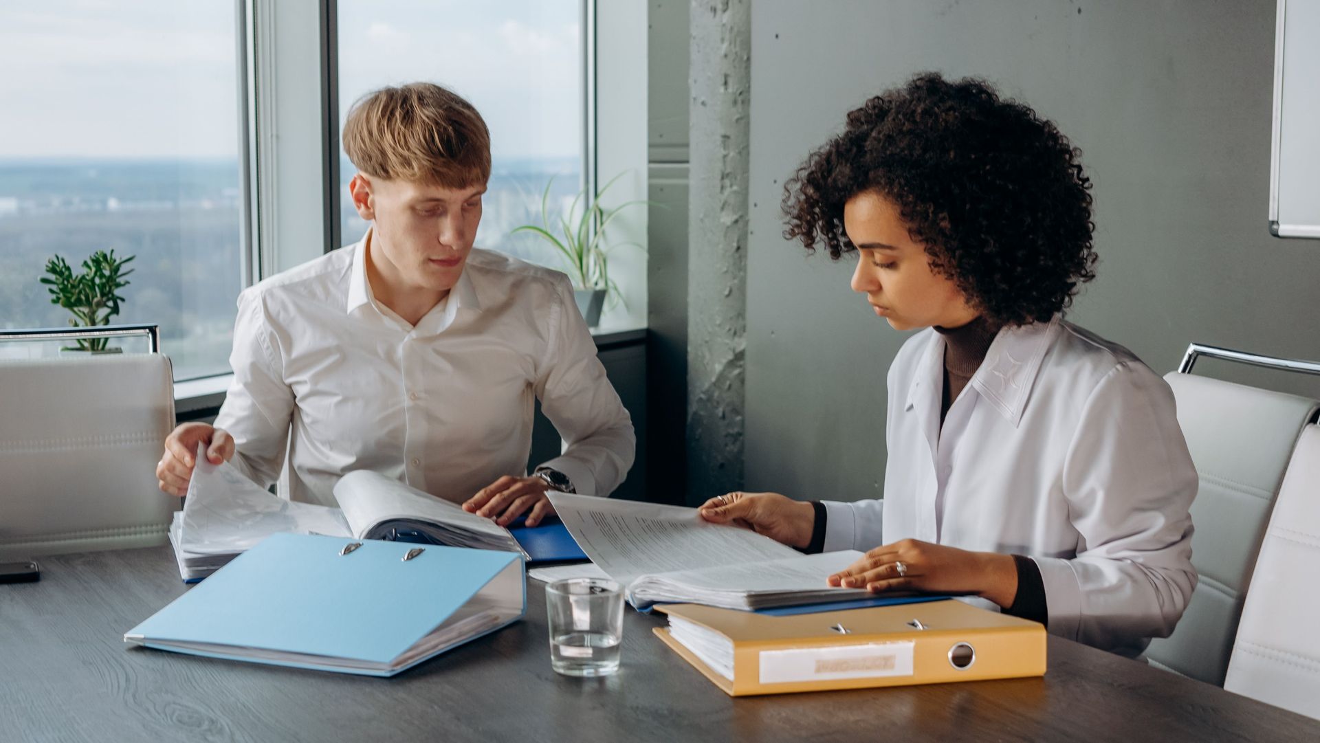 Two coworkers review papers at a table, with folders and a glass of water in a bright office.