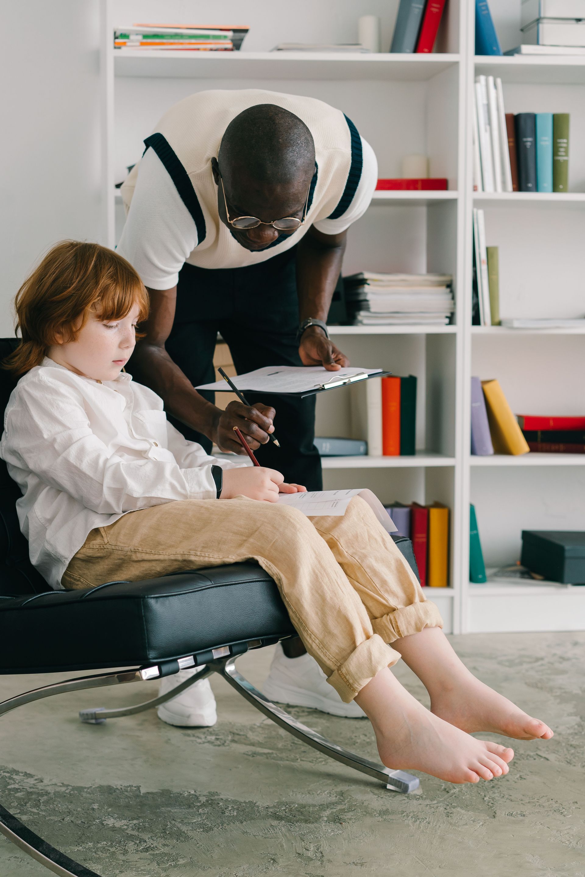 Adult helping child read in a bright living room with white shelves and a black chair