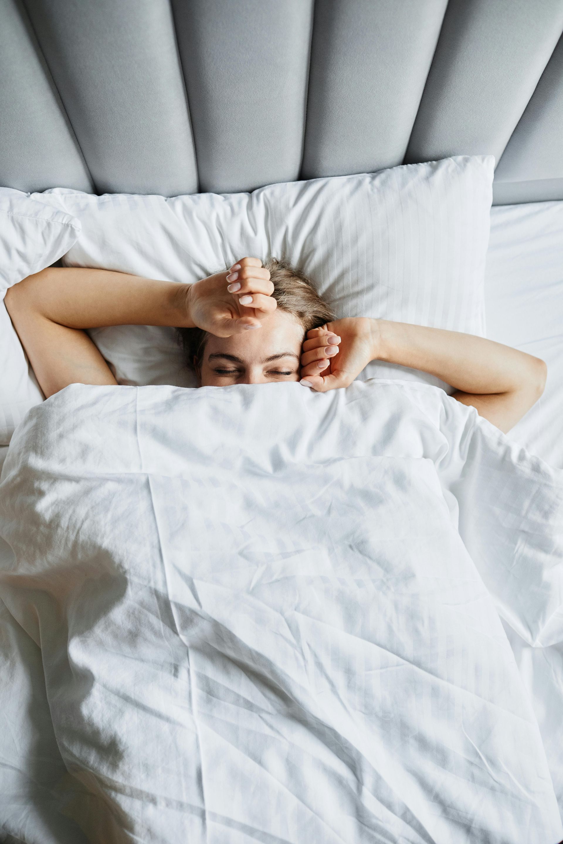Person lying in bed under white sheets with hands on forehead, resting in a bright bedroom