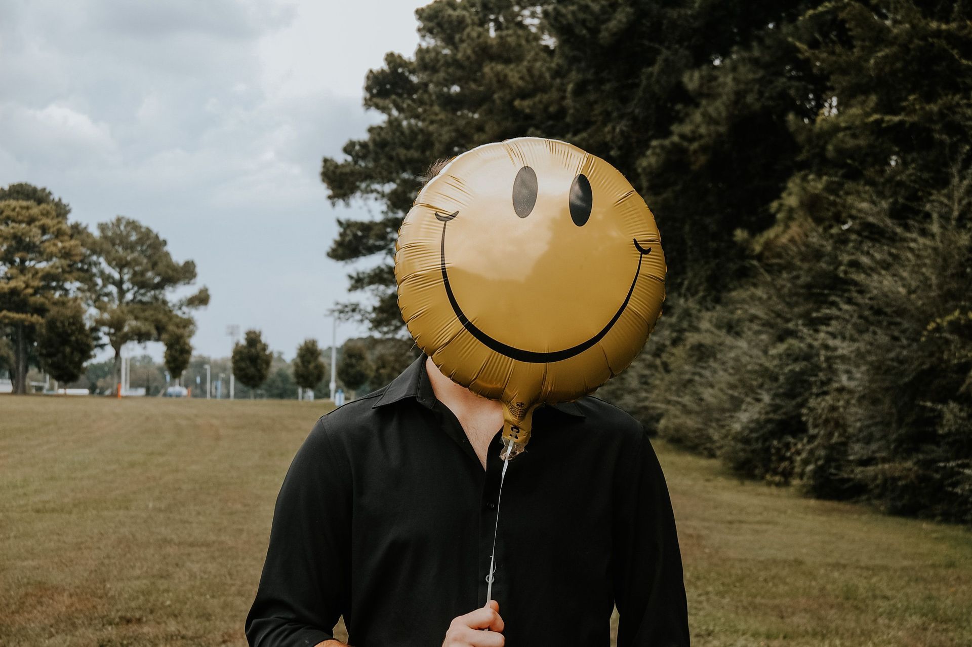 Person holding a large yellow smiley-face balloon in a grassy field with trees in the background