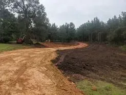 Dirt road and excavated land with trees in the background under a cloudy sky.