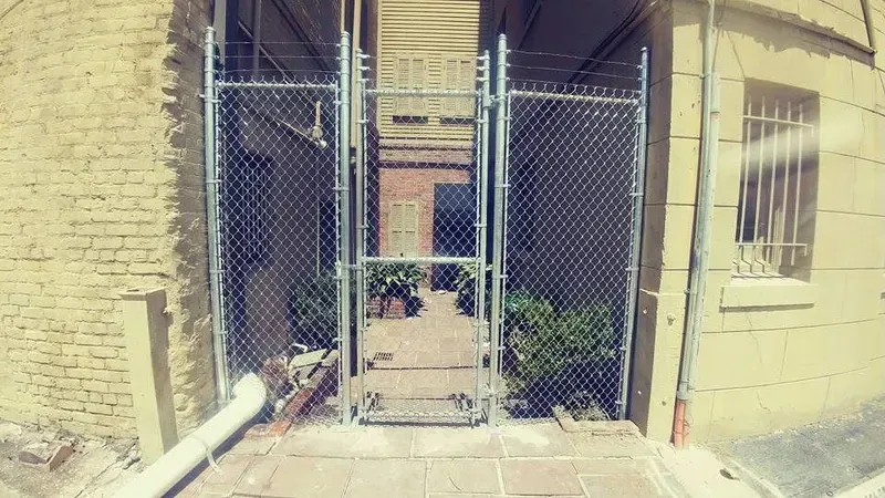 Chain-link fence gate opening to a brick courtyard with buildings and plants, with sunlight.