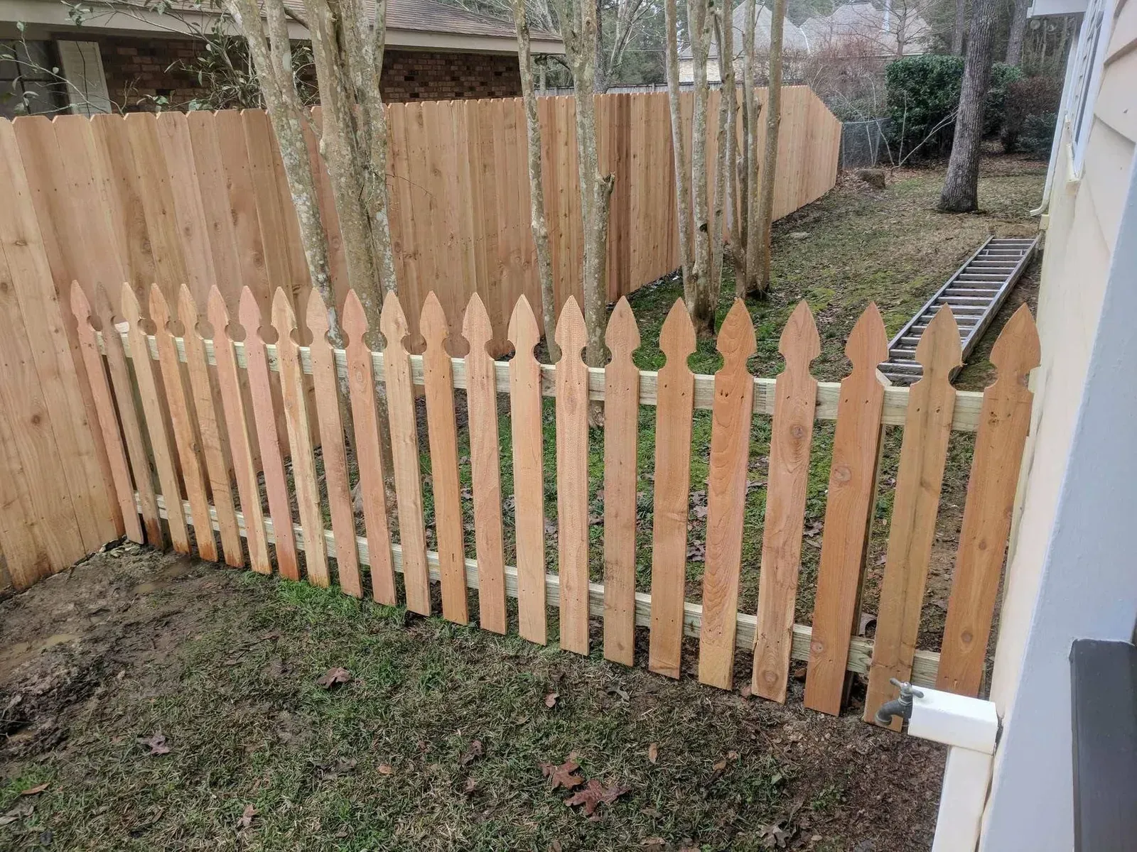 Wooden picket fence in front of a tall wooden privacy fence in a backyard.