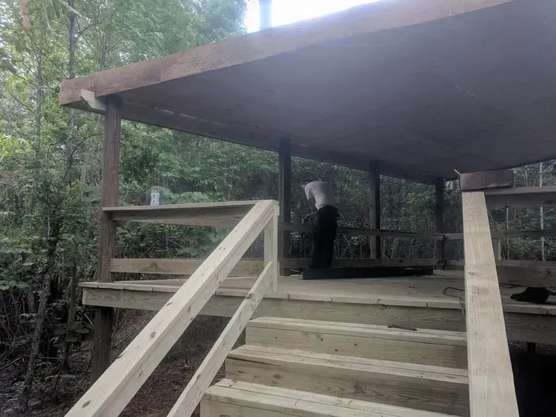 Wooden deck and steps leading to a covered structure in a wooded area. Person standing inside.