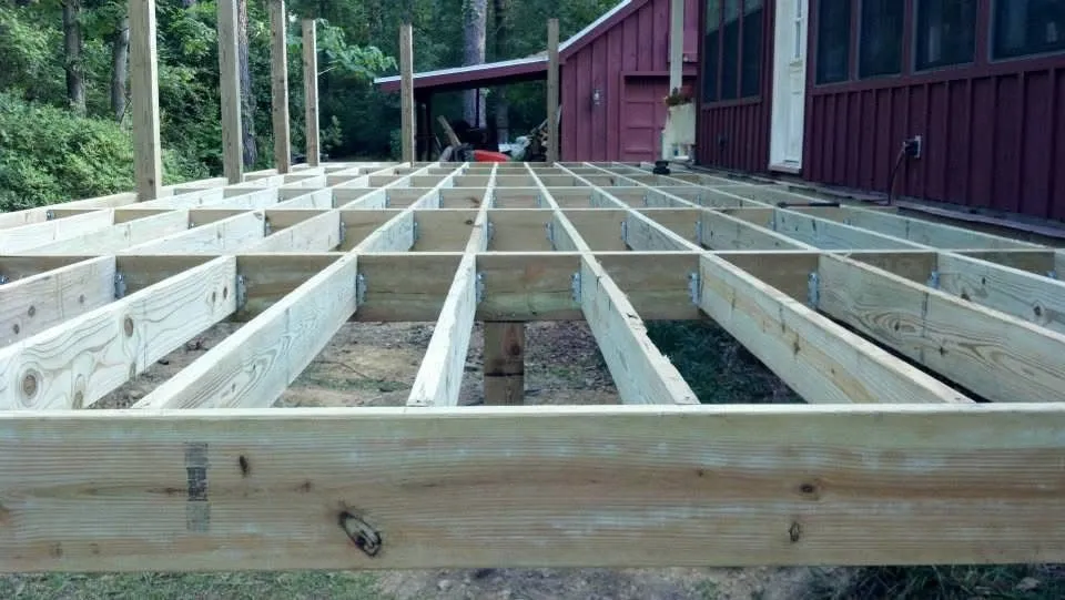 Wooden deck frame under construction next to a red building; trees in background.
