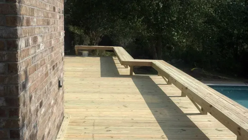 Wooden deck with built-in bench next to a brick wall and a pool.