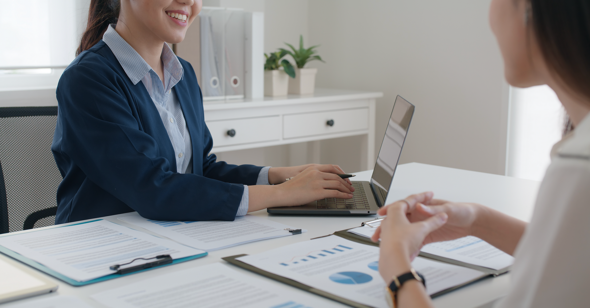 A woman is sitting at a desk with a laptop and talking to another woman.