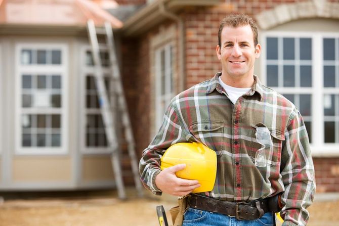 Man standing outside a house, holding a yellow hard hat and smiling at the camera.