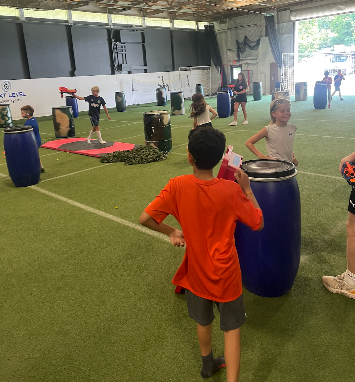 A boy in an orange shirt stands next to a blue barrel
