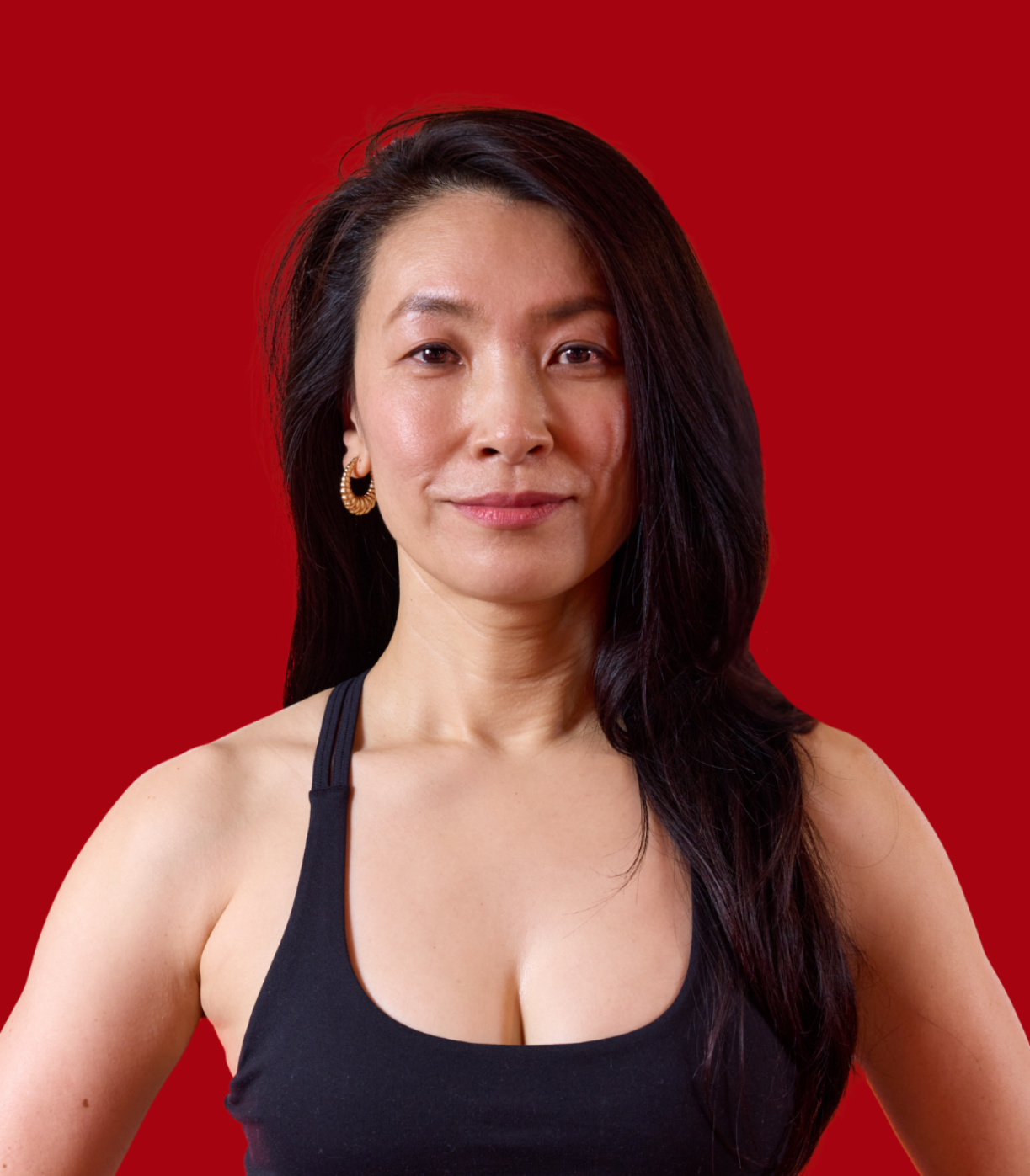 Closeup of a smiling woman with long dark hair in yoga clothing with a medium-red background.