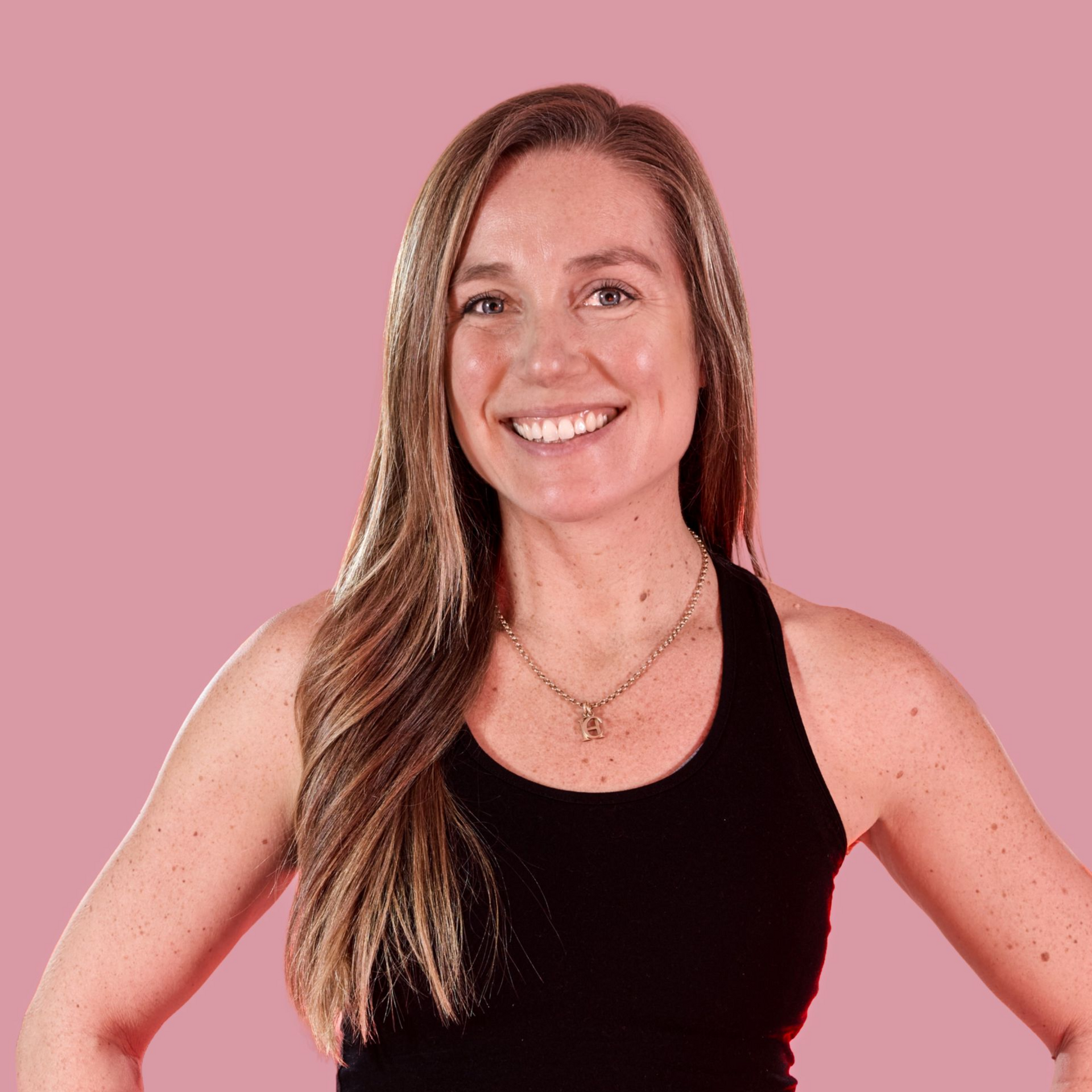 Closeup of a smiling woman with long blonde hair in a black yoga set with a muted pink background.
