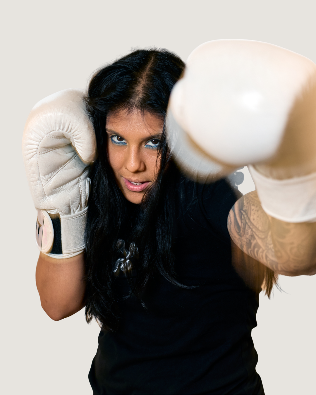 Closeup of a woman with long dark hair and boxing gloves in front of an off-white background.
