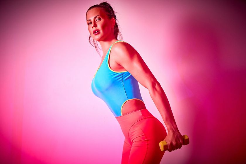 Woman in colorful workout clothes holding weights in a room with bright pink lighting.