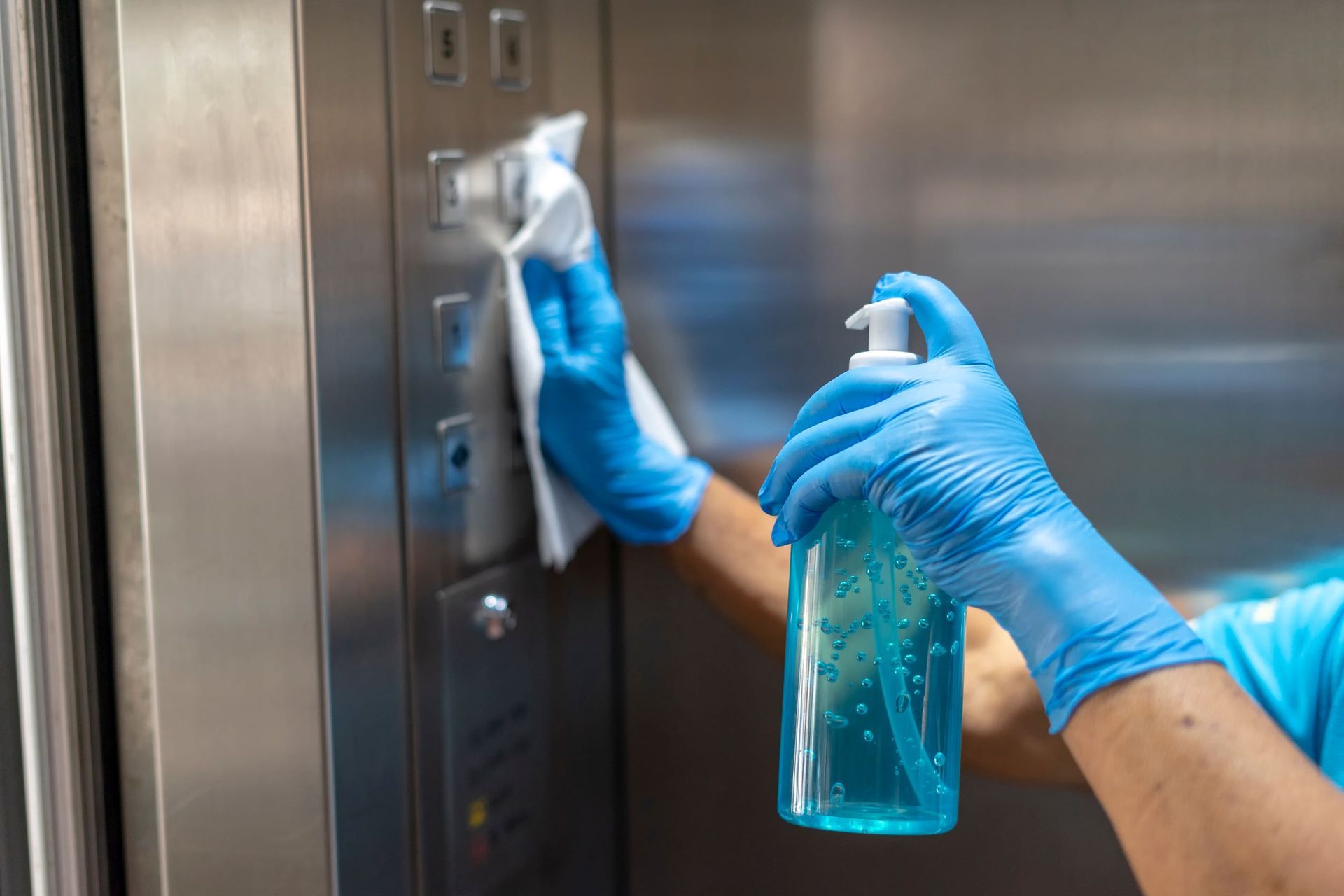 Gloved hands wiping a metal elevator panel with blue cleaning spray.