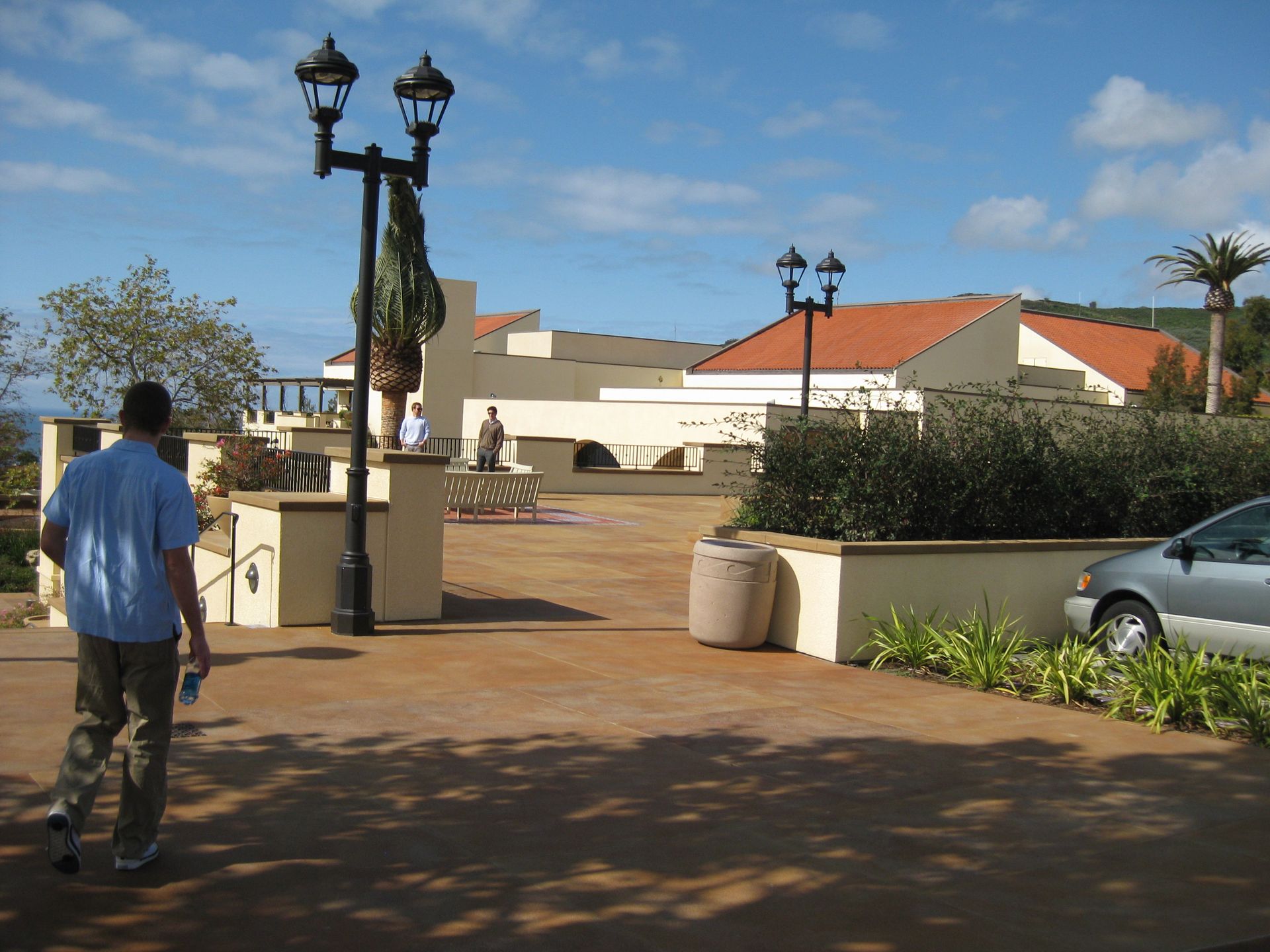 Person walking on a sunny plaza near white buildings and a parked car.