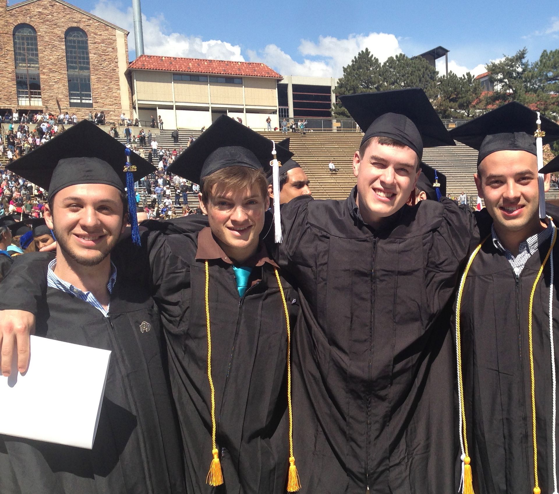 Four graduates in caps and gowns smiling outdoors at a commencement ceremony