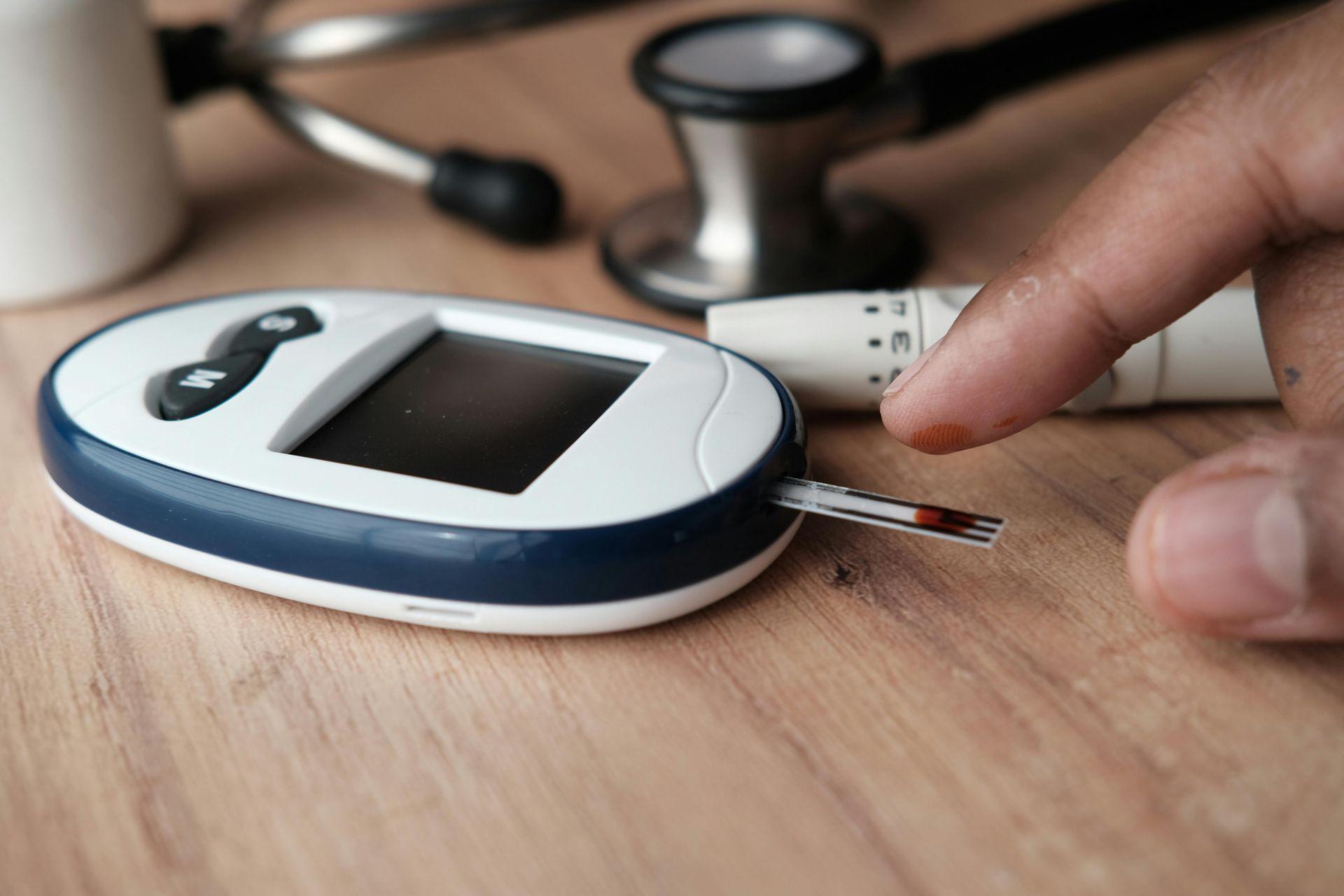 Hand pricking finger for blood glucose test; meter, lancet, stethoscope on wooden table.