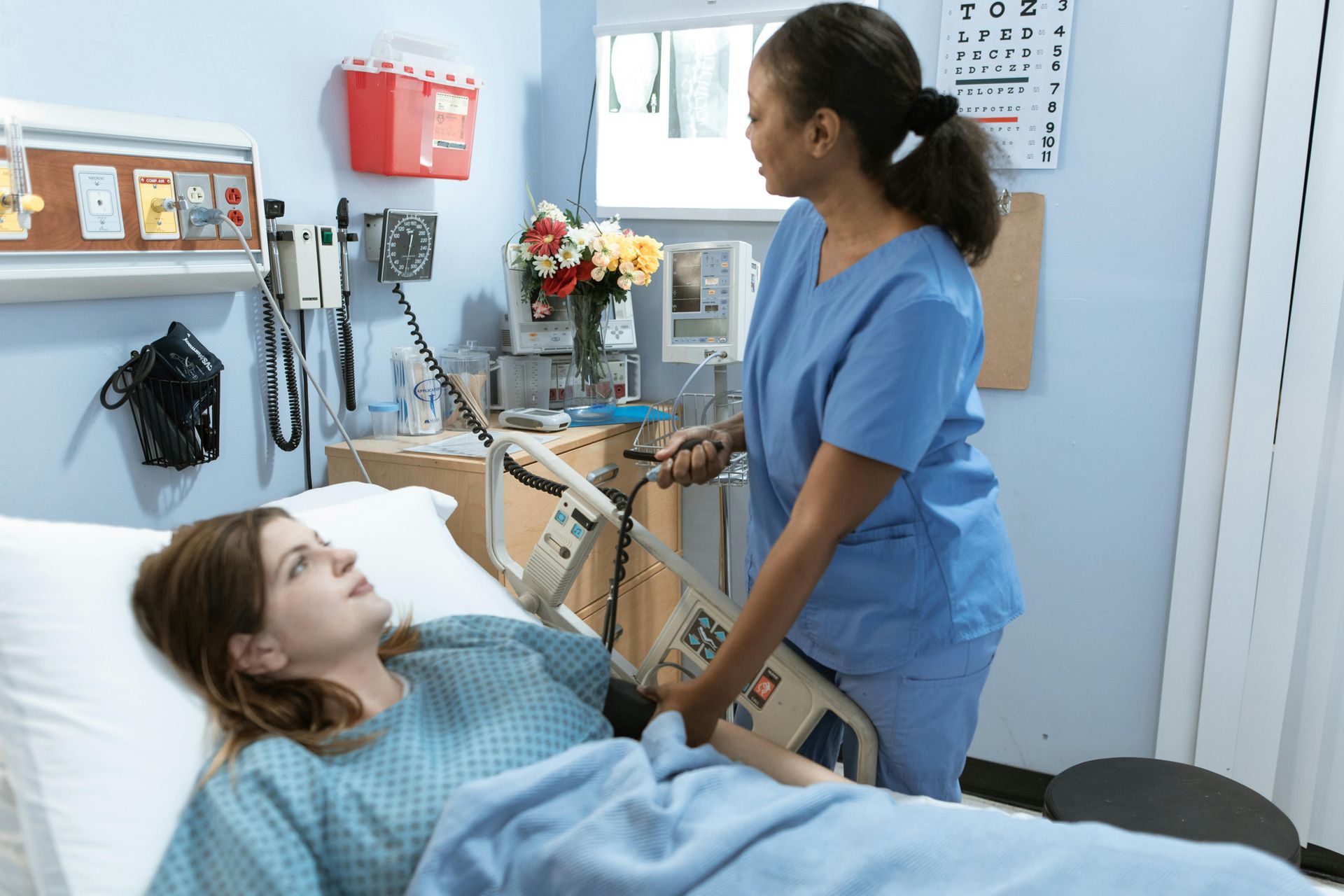 Nurse in blue scrubs attending to a woman in a hospital bed; medical equipment in the room.