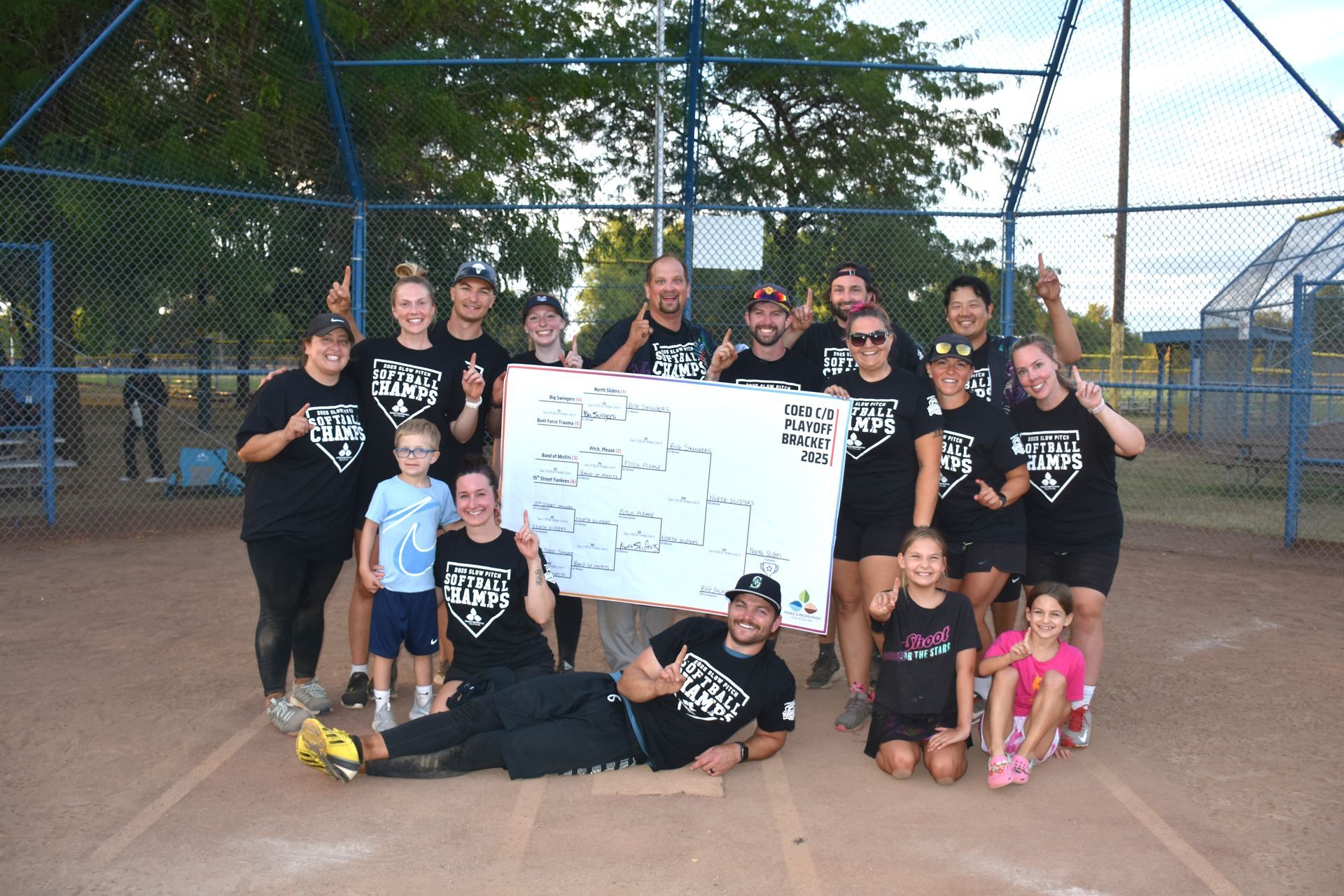 A softball team wearing matching black shirts poses for a photo on a dirt field, holding a tournament bracket poster.