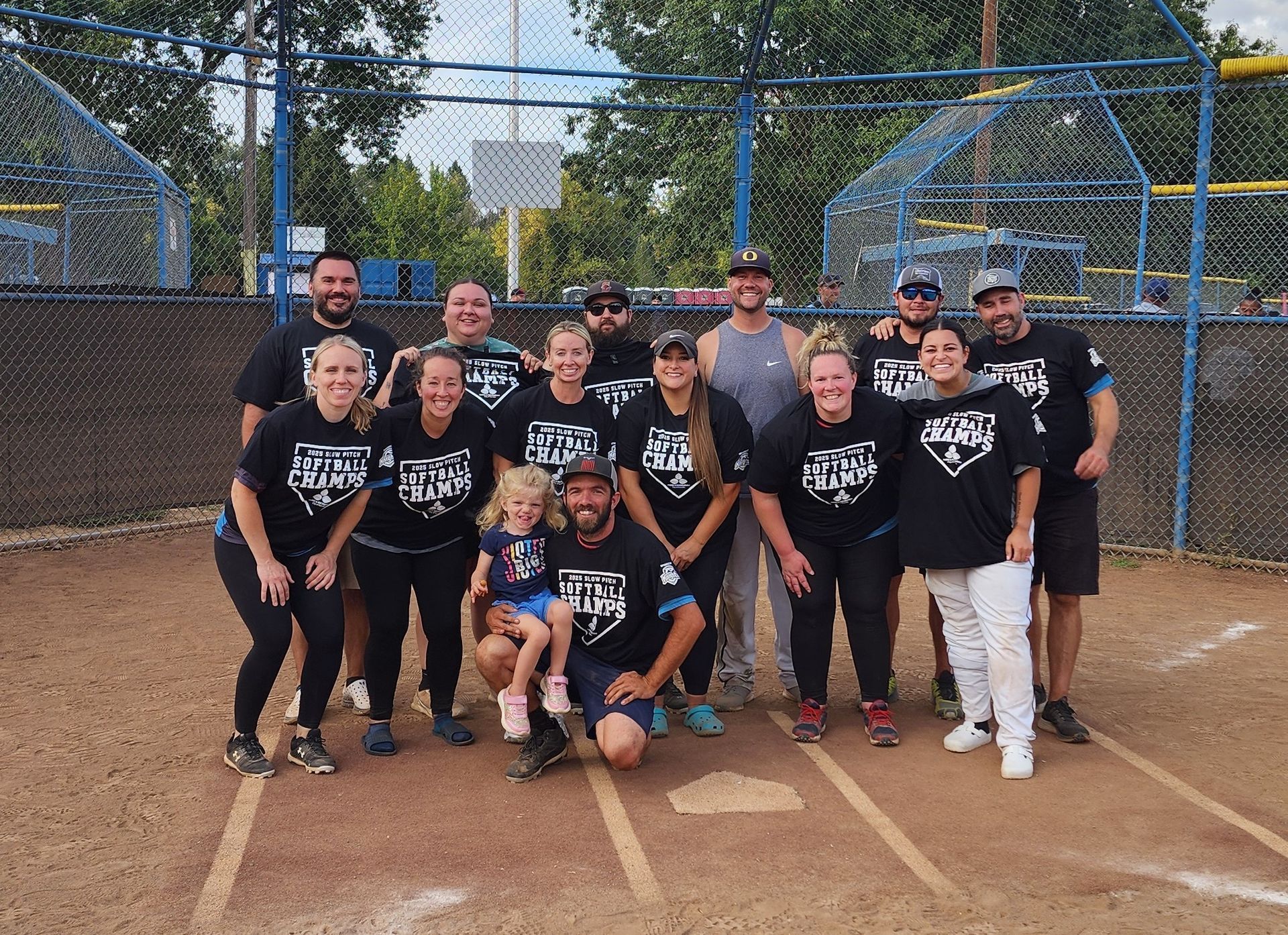 A men's softball team wearing matching black shirts poses for a photo on a dirt field in front of a chain-link backstop.