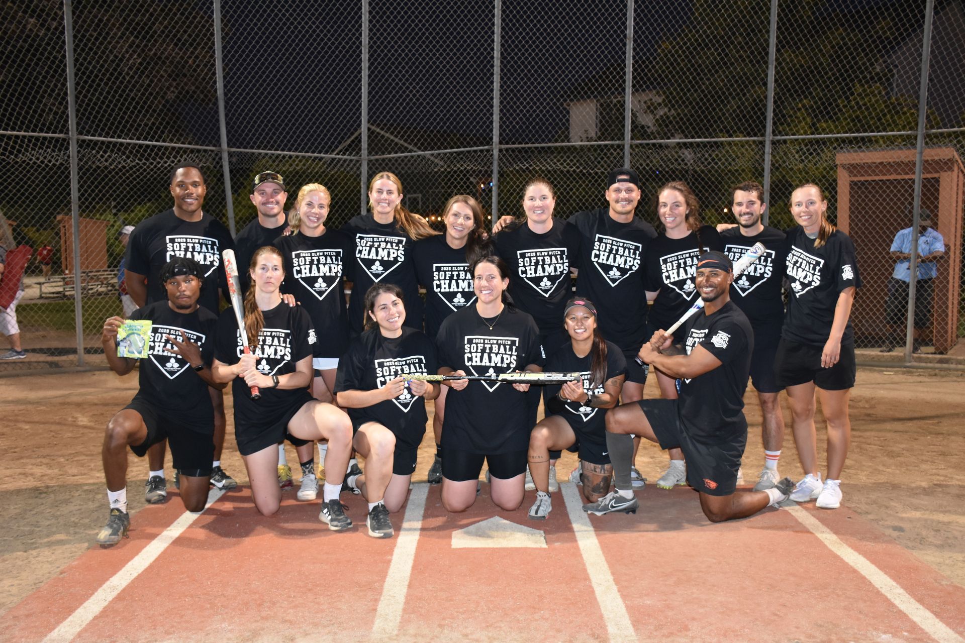A softball team poses for a group photo on a dirt field at night, wearing matching black shirts and holding bats.