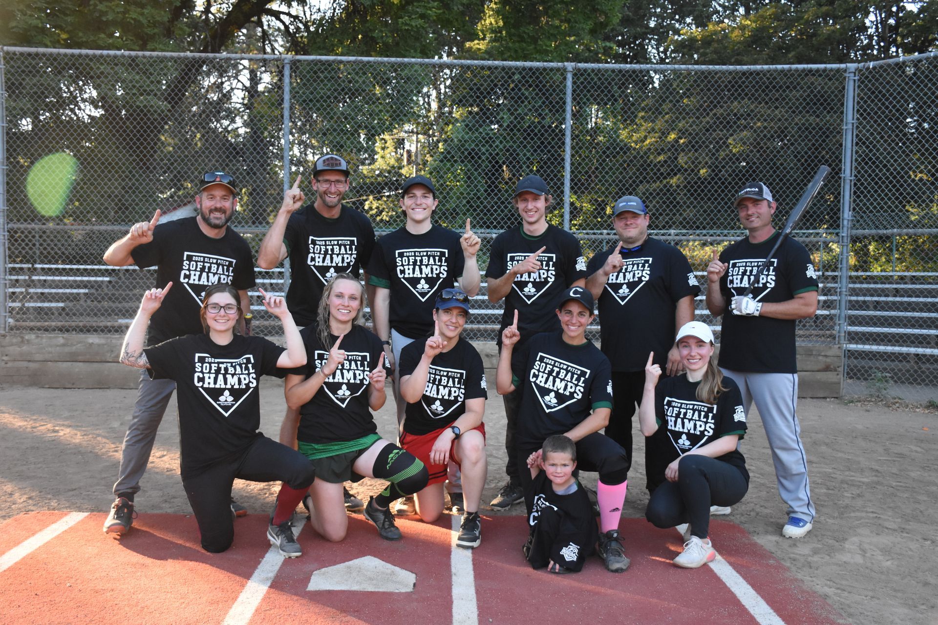 A softball team in matching black t-shirts and baseball caps posing on a dirt softball field after a championship win.