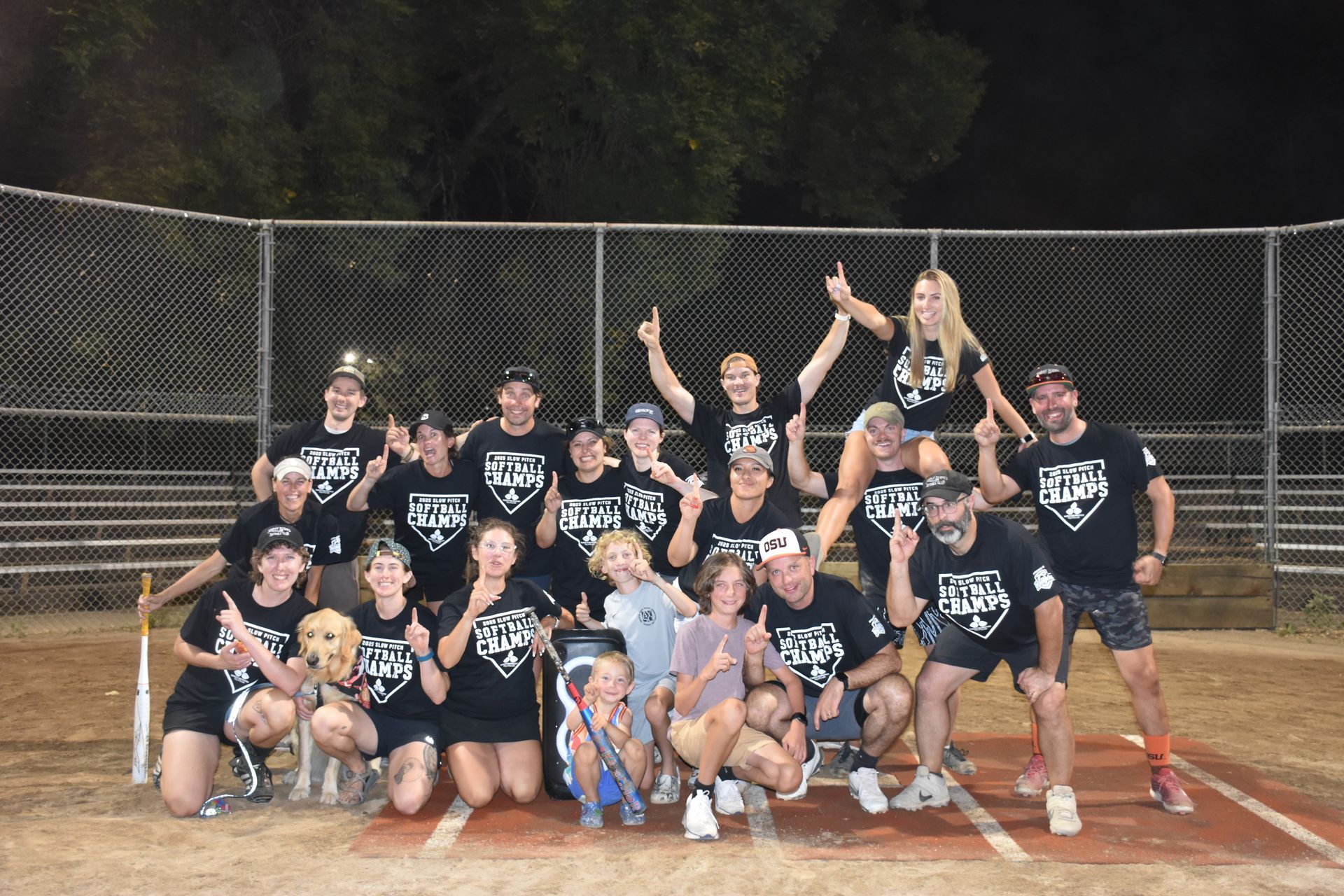 A softball team in matching black shirts poses for a group photo on a baseball field at night.