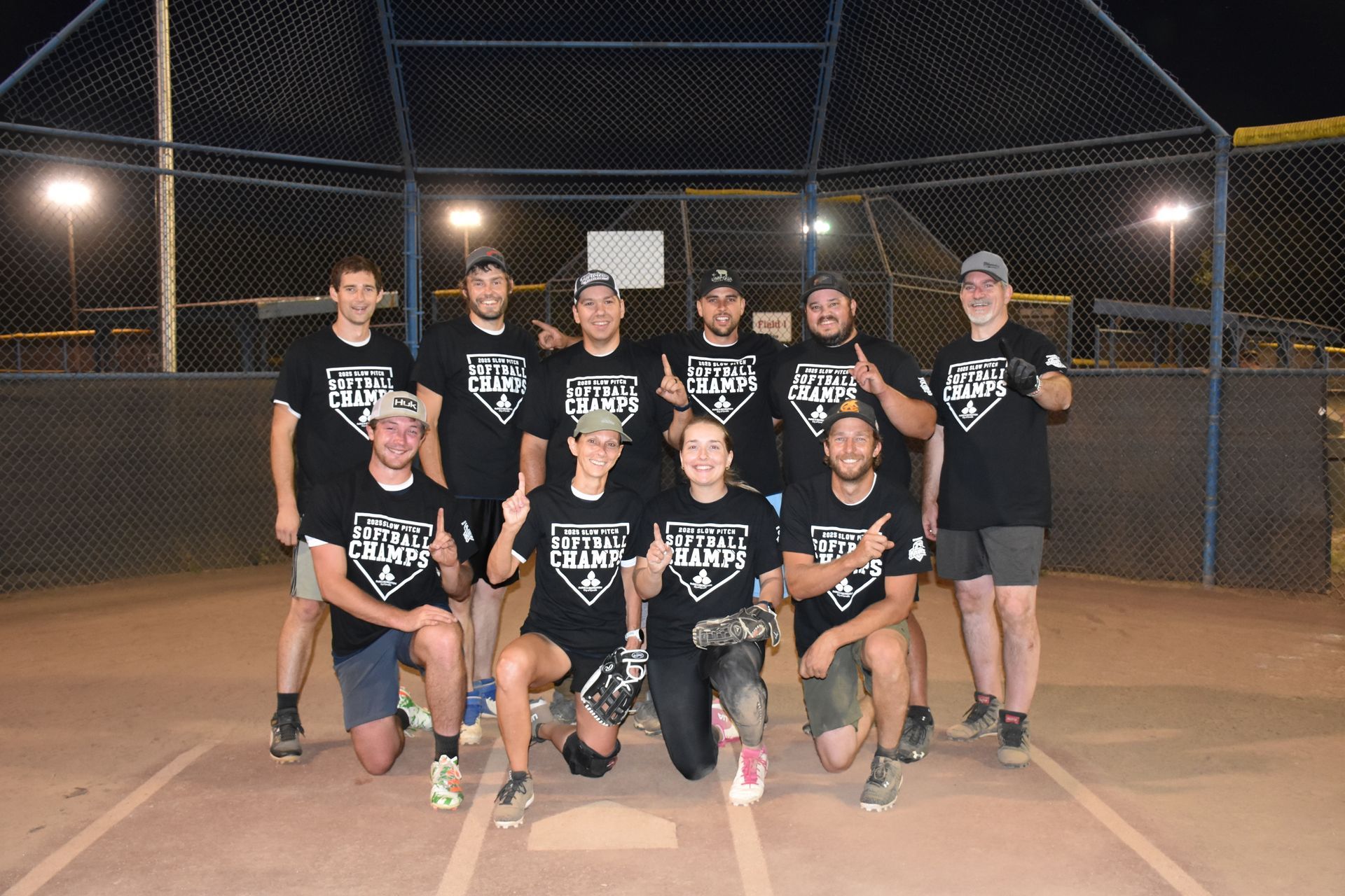 A softball team wearing matching black shirts poses for a photo on a dirt field at night under bright stadium lights.