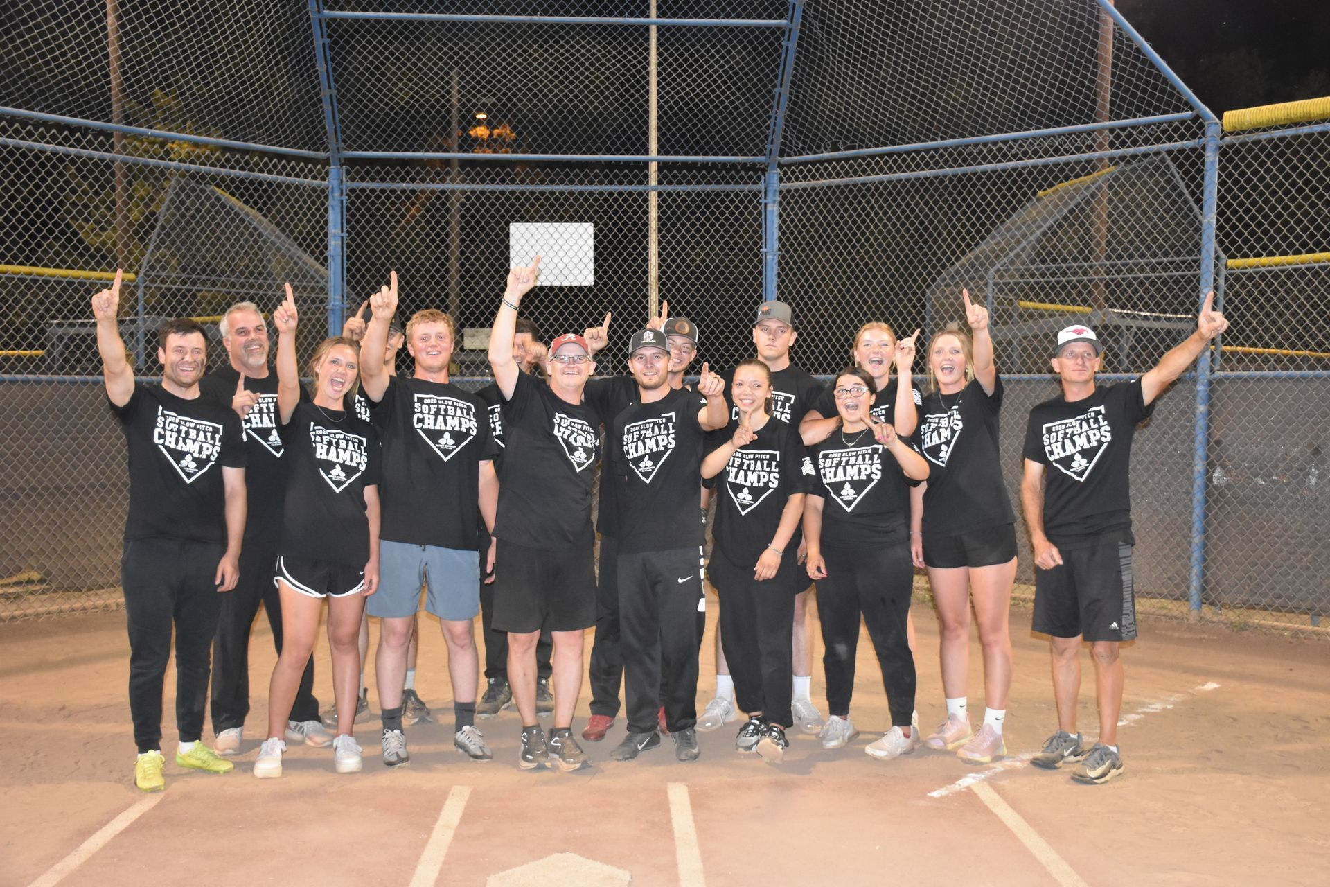 A softball team in black shirts stands on a field at night, holding up index fingers in celebration.