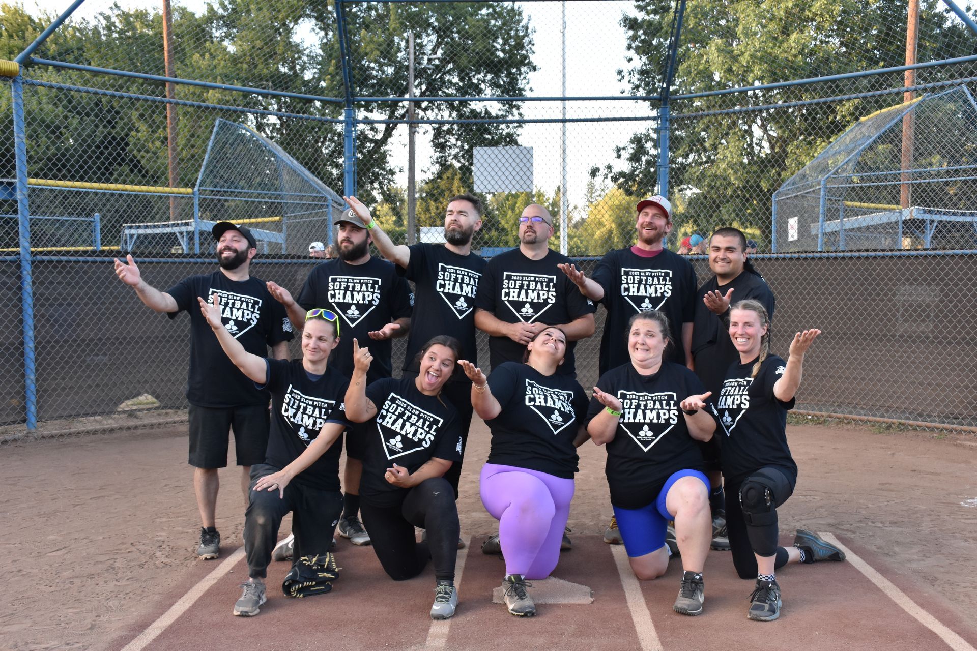 A softball team in matching black shirts poses on a baseball field, most looking upward with arms extended toward the sky.