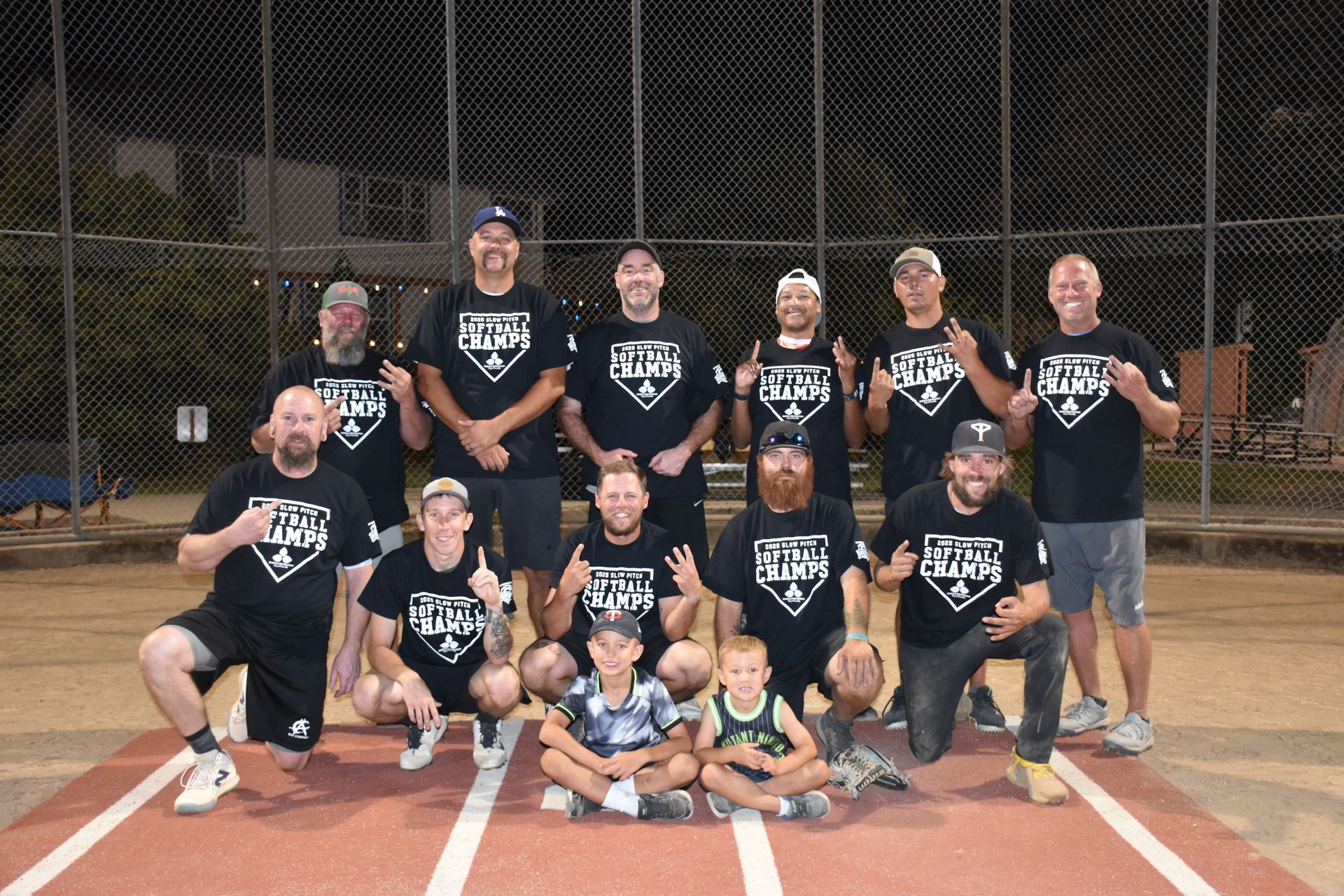 A group wearing matching shirts poses on a baseball field at night, some kneeling with children in front of them.