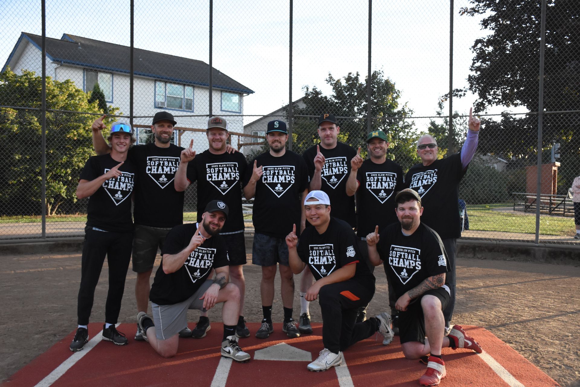 A softball team in matching black shirts celebrates a win on a dirt field, with some players gesturing one finger up.