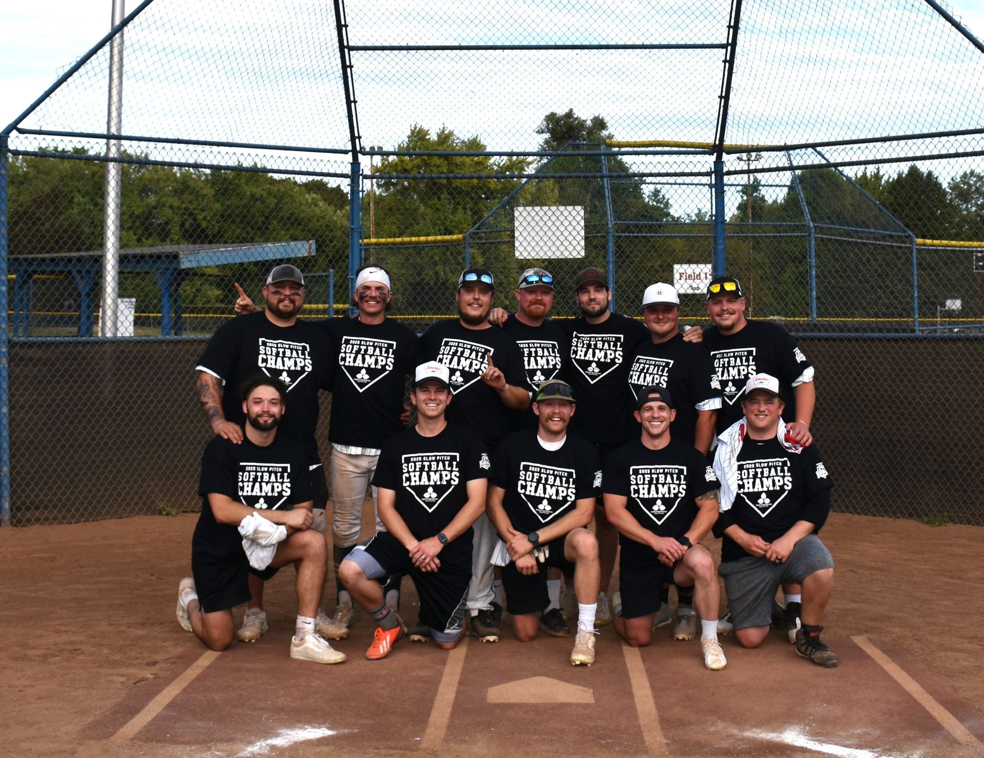 A men's softball team wearing matching black shirts poses for a photo on a dirt field in front of a chain-link backstop.