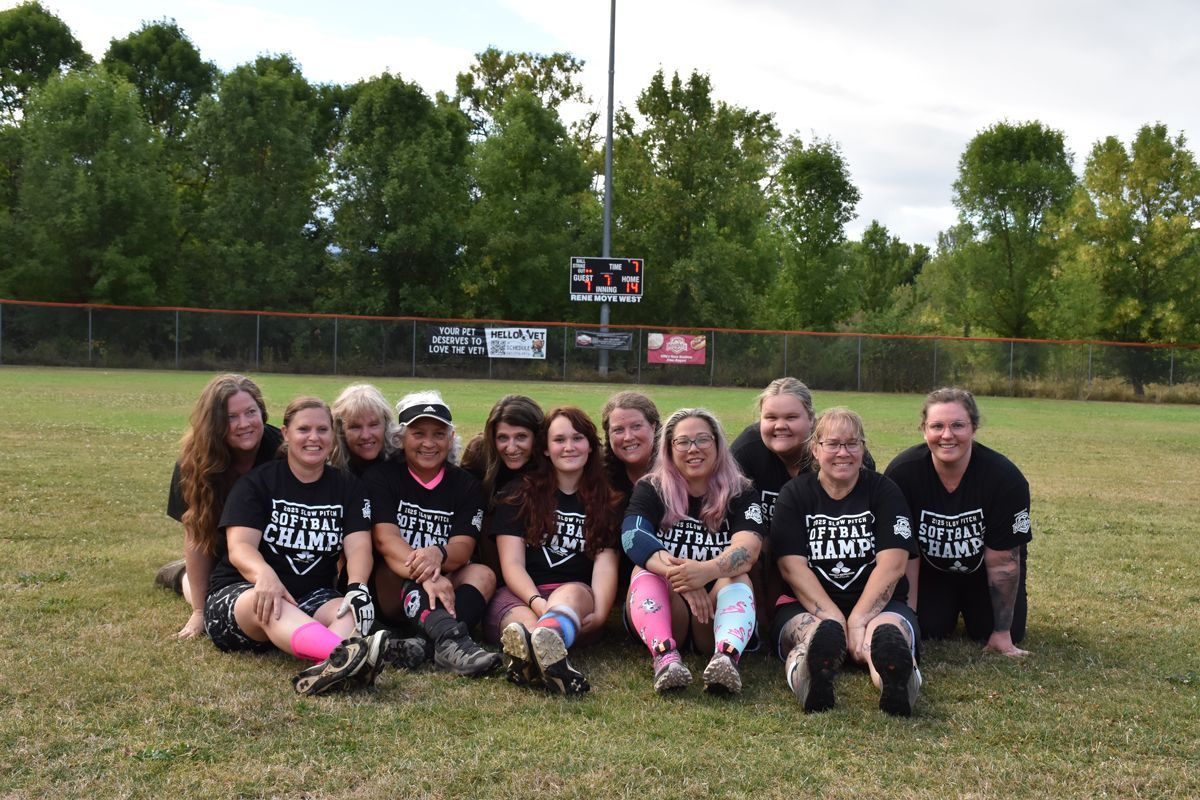 A team of twelve people wearing black matching t-shirts pose together on a grass field in front of a baseball fence.