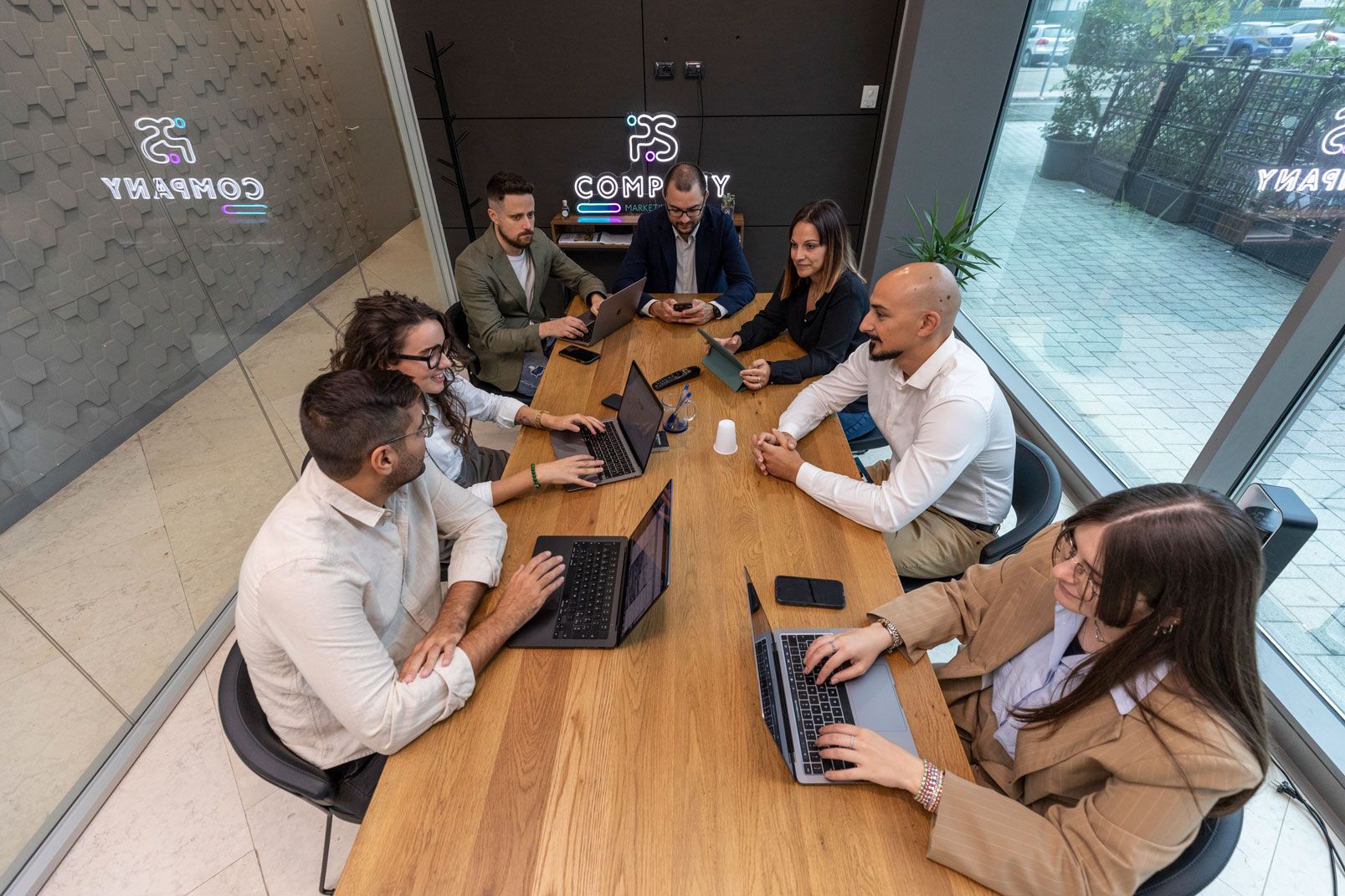 Un gruppo di sette persone sedute a un tavolo di legno in un ufficio, lavorano con i computer portatili.