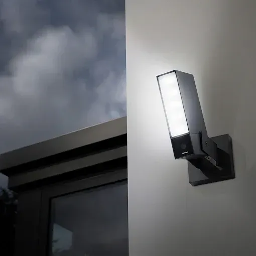 Mani che reggono uno smartphone mostrando i comandi della domotica davanti a una casa moderna al crepuscolo.