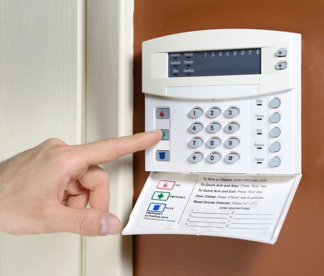 Una mano che regge uno smartphone con un'app per la sicurezza domestica davanti a una moderna casa bianca.
