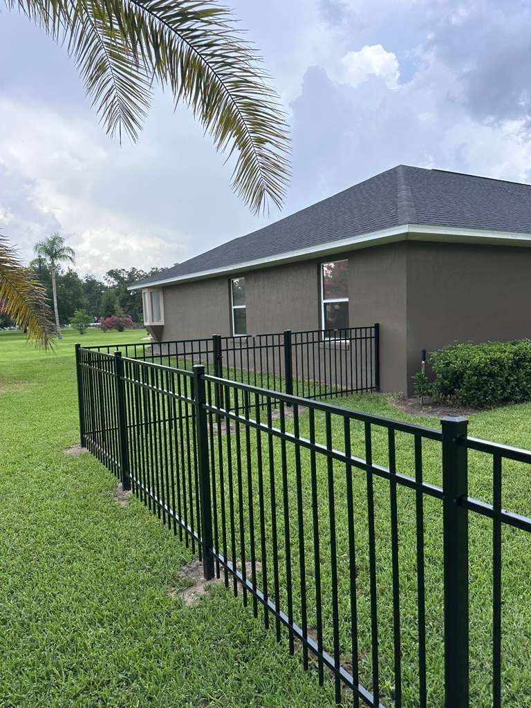 Black metal fence beside a gray house on a green lawn under cloudy sky