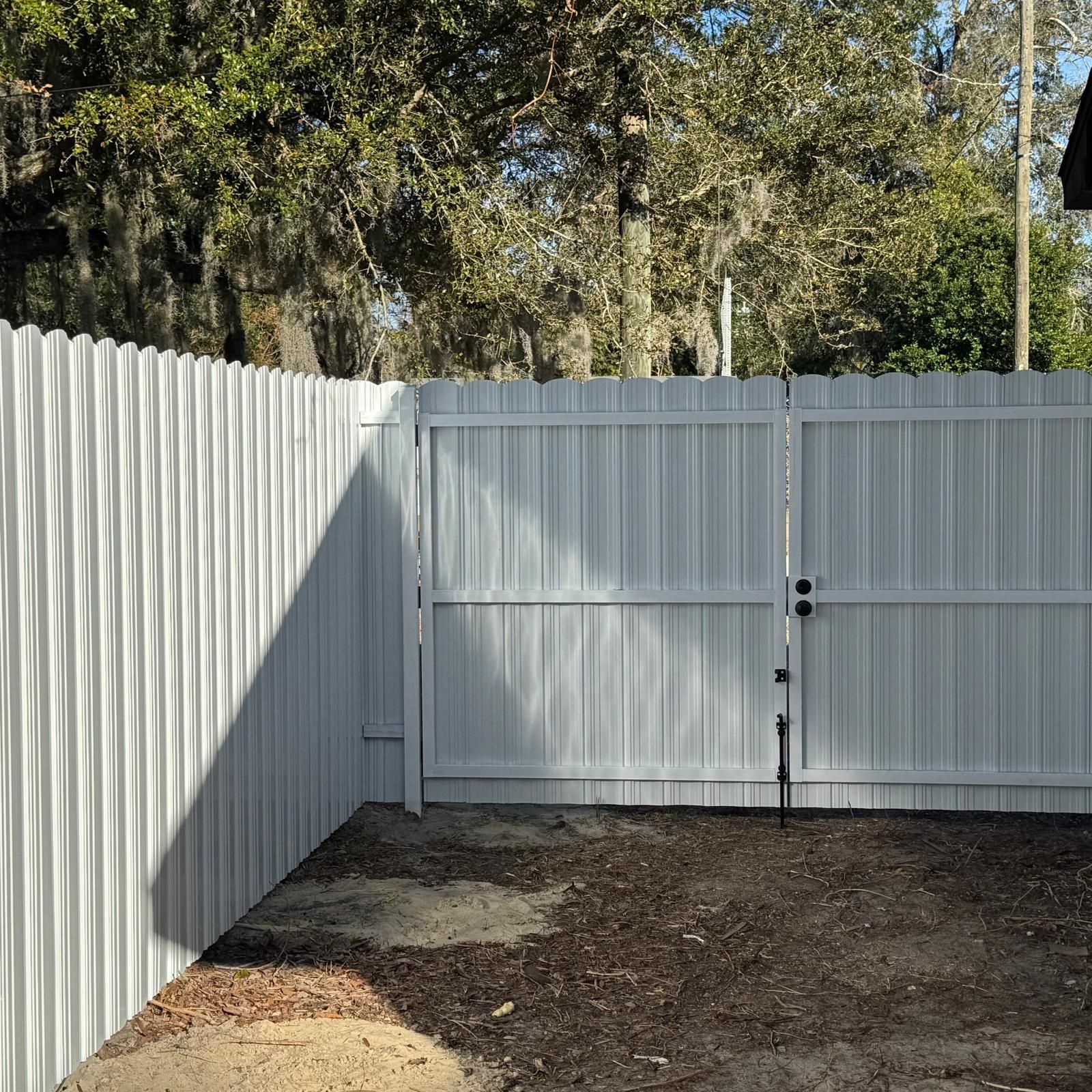 White metal fence and gate enclosing a dirt yard with trees behind it
