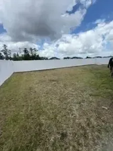 Grassy field beside a white wall under a cloudy sky, with trees in the distance.