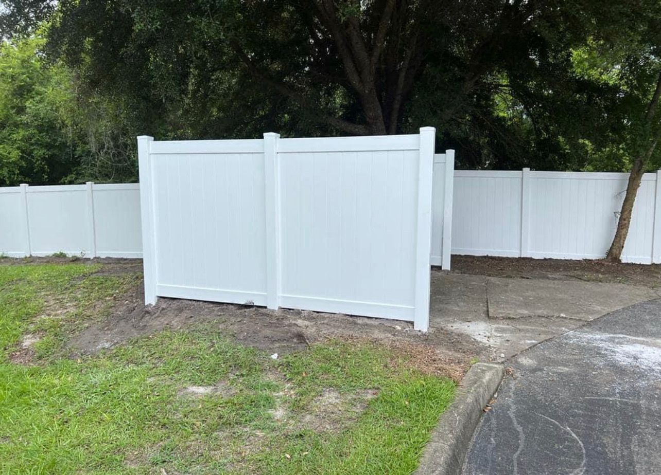White vinyl double gate in a fenced yard beside a paved driveway, with trees and grass in the background