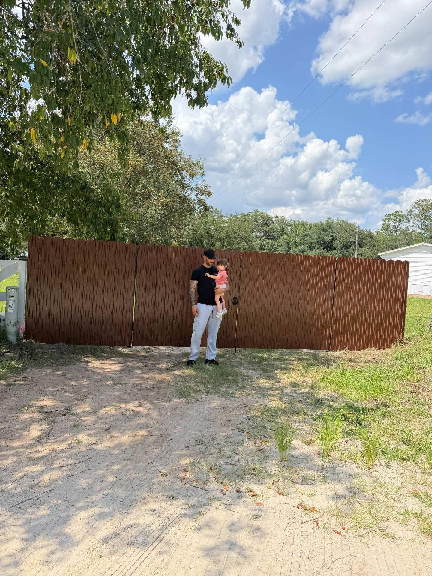 Two people standing in front of a large brown fence on a sunny dirt lot with trees and clouds overhead