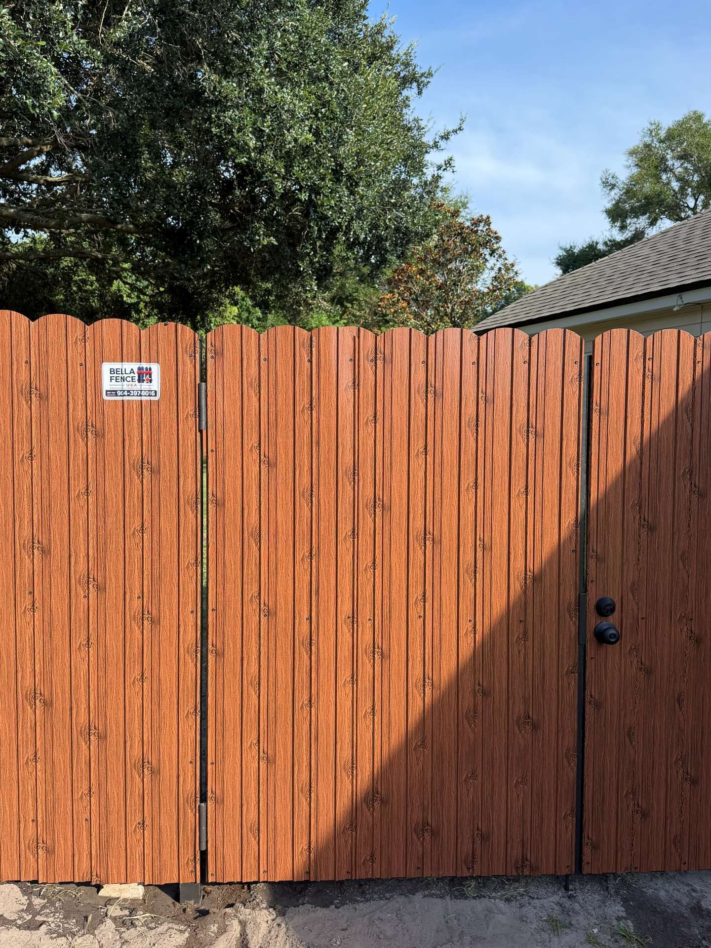 Tall orange wooden fence with a small sign, trees above, and a gate in sunlight and shadow