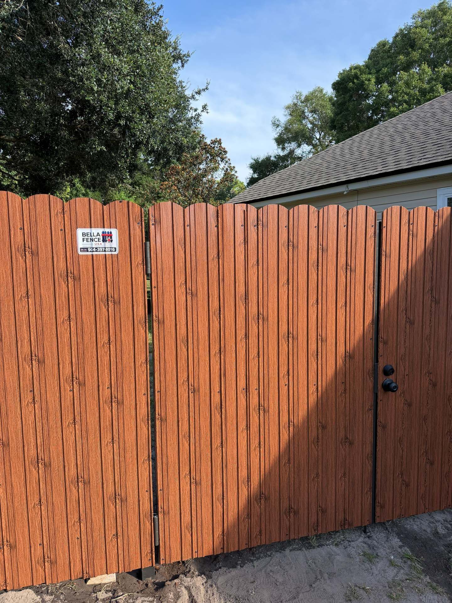 Wooden privacy fence and gate with a small sign, trees above, and a concrete driveway in front.
