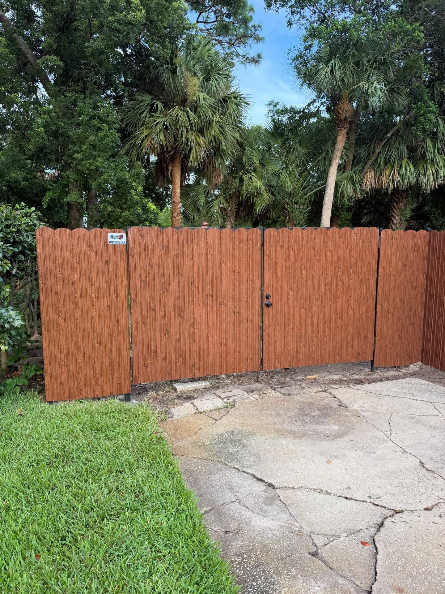 Wooden fence with a small gate beside a cracked concrete driveway and green lawn, with trees behind it