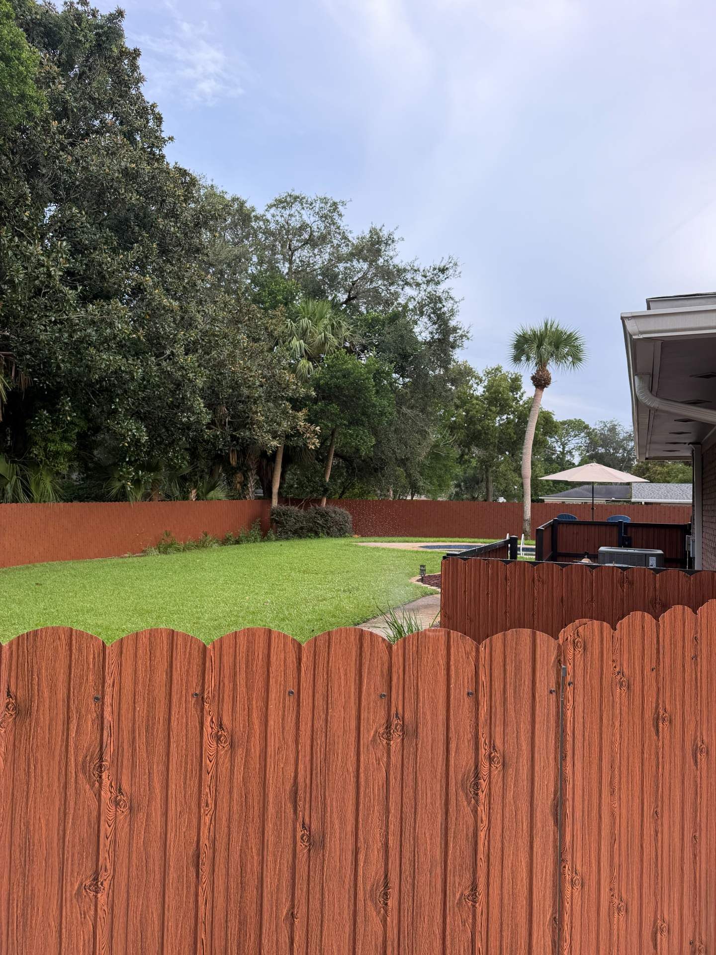Backyard with green lawn, red fence, trees, and a small patio under a cloudy sky