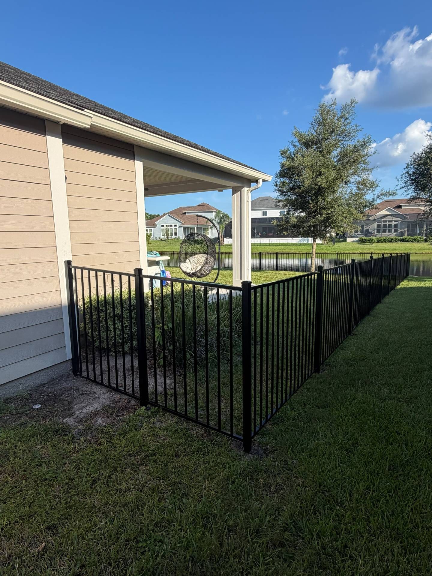 Black metal fence beside a beige house, bordering a grassy yard under a blue sky