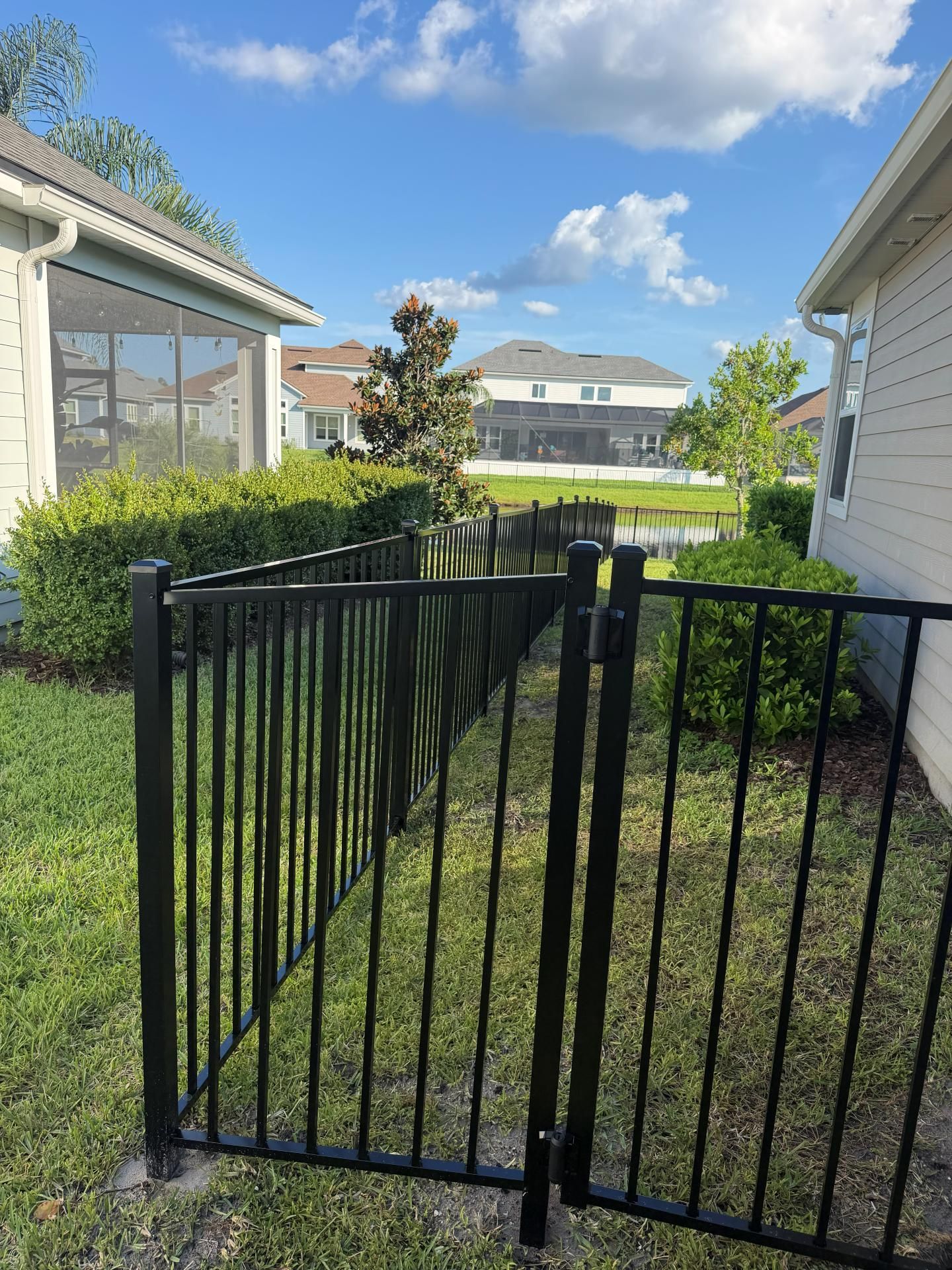 Black metal gate and grassy side yard between houses under a partly cloudy blue sky.