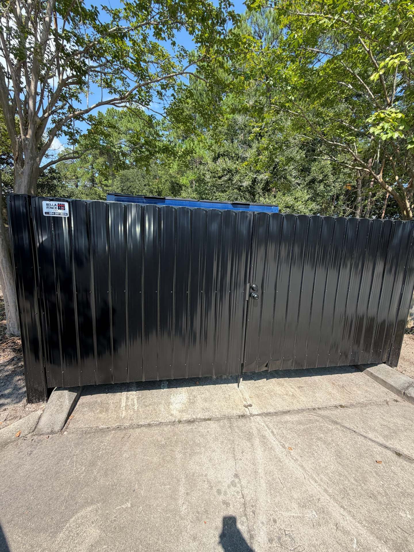 Black corrugated metal fence panels along a sidewalk beside trees and concrete pavement.
